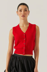 A woman is wearing a sleeveless red knit vest with a V-neckline and button-down front. The vest has a fitted ribbed hem at the waist, creating a cropped look. She is paired with black, loose, gathered pants.